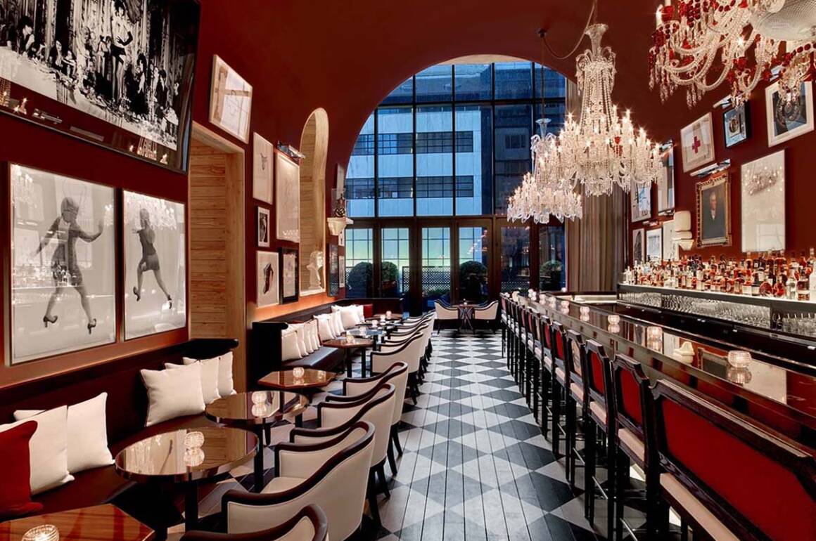 A red restaurant with a large window at the back and long rows of tables with benches and chairs on either side and crystal chandeliers over the bar