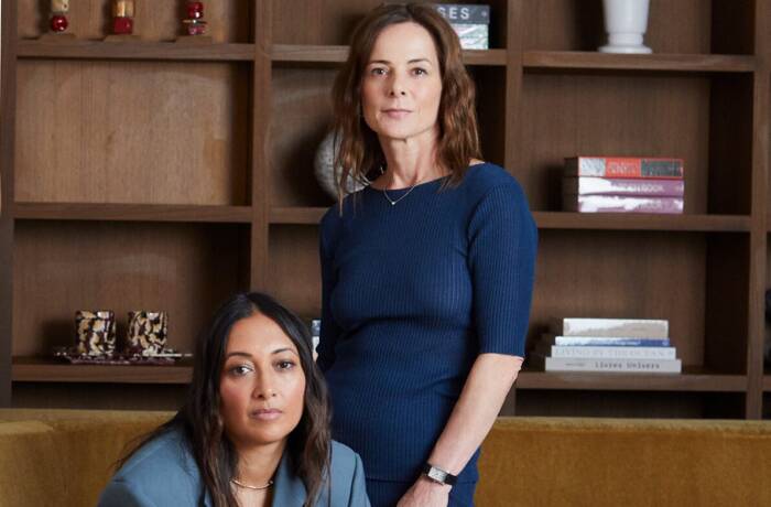 two women, one sitting one standing, wearing blue with a brown background